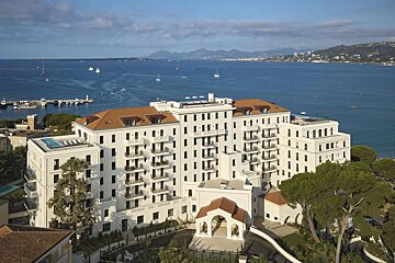 An elegant white hotel with red roofs and balconies sits above a sparkling blue bay dotted with boats and a distant coastal city.
