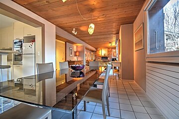 An inviting, long dining area with a glass table and chairs, featuring a warm wooden ceiling, leading to an open kitchen and a cozy living room.