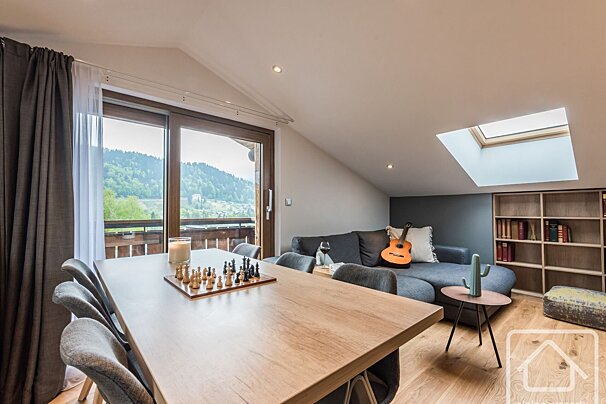 Bright, modern chalet living/dining room with a mountain view balcony. Chess set on table, cozy sofa, guitar, skylight, and bookcase.