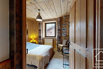 A cozy, rustic bedroom with wood-paneled walls and ceiling, a bed, and a window. Alpine-style decor includes patterned curtains and animal art.