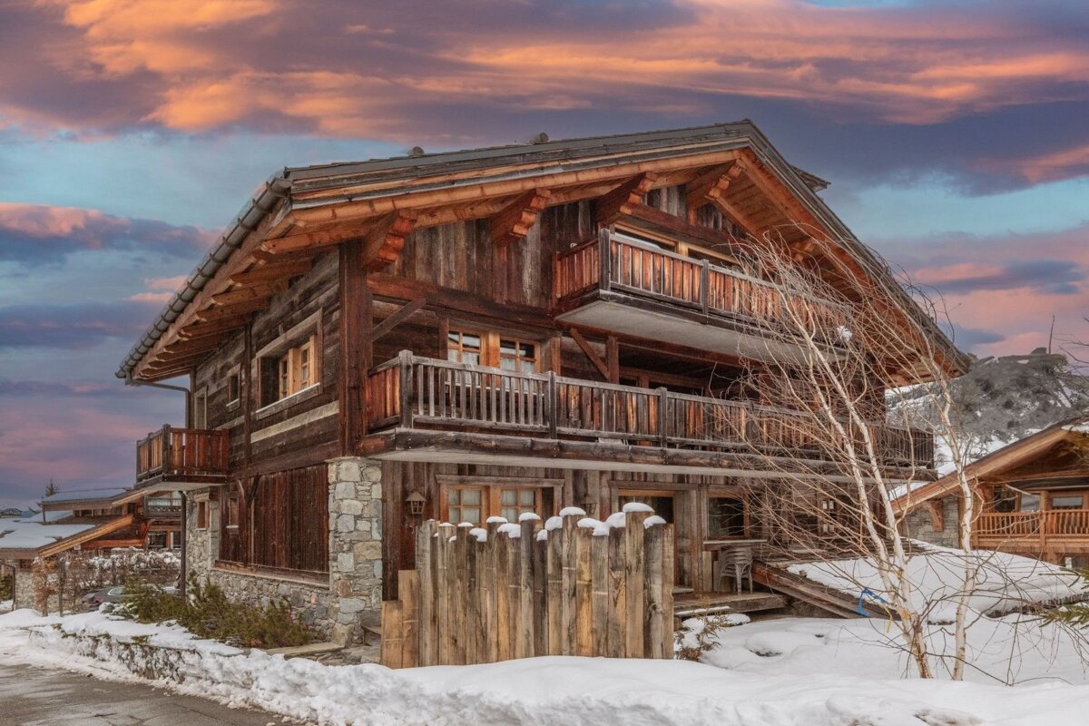 A rustic wooden chalet with balconies stands in a snowy mountain landscape under a vibrant pink and blue sunset sky.