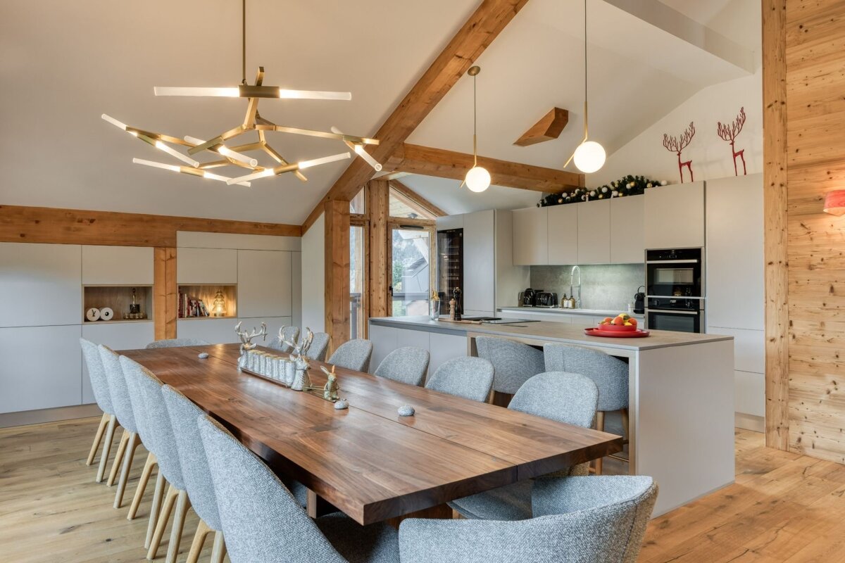 A spacious modern rustic kitchen-dining room with wooden beams, sleek grey cabinets, a long table, contemporary lighting, and festive deer decorations.