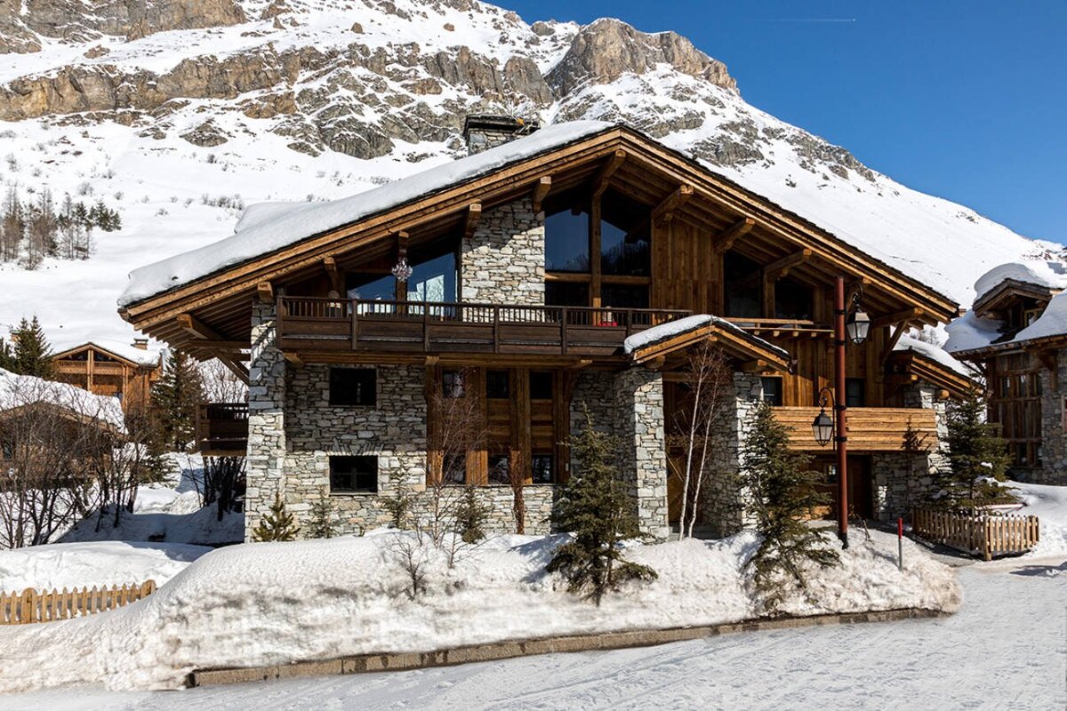 A luxurious stone and timber chalet with a snowy roof, nestled in a snow-covered mountain landscape under a clear blue sky.