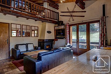 Cozy chalet living room with a fireplace, leather sofas, wooden beams, ski decor, a loft, and a view of a green outdoor area.