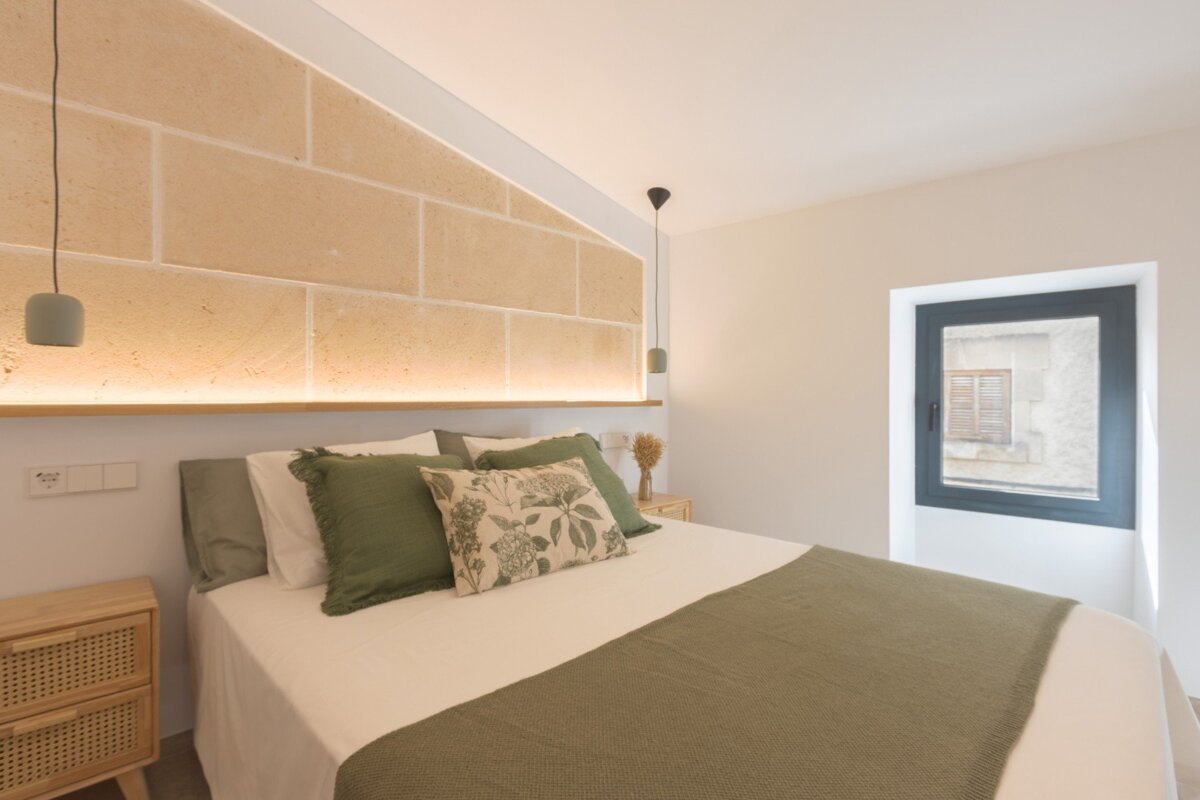 Cozy bedroom with a bed featuring green linens, a lit stone accent wall, and a dark-framed window.