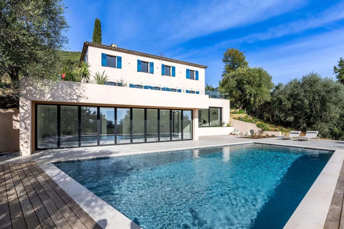 A modern white house with blue shutters, a glass-walled lower floor, and a large swimming pool, set against a sunny, tree-covered hillside.