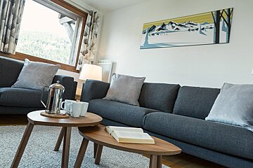 A modern living room with grey sofas, nested wooden coffee tables with a coffee press and book, and a large window overlooking a forest. Mountain wall art completes the decor.