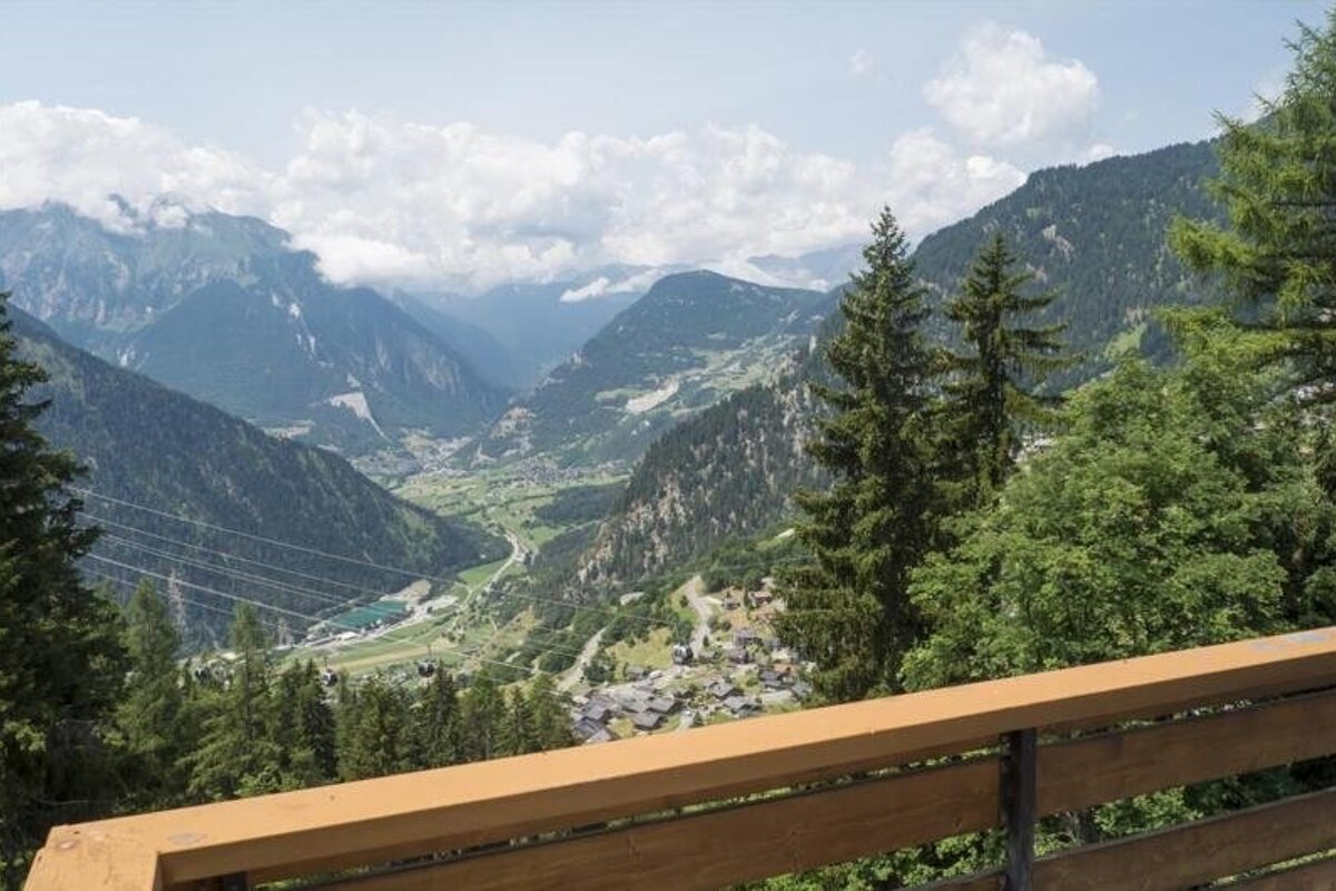 Scenic view of a lush green alpine valley with mountains and pine trees, taken from a wooden balcony.