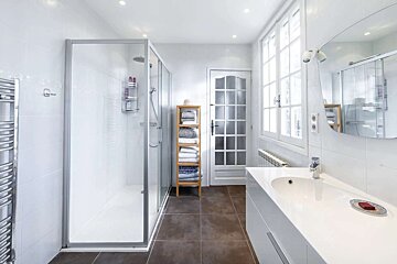 Bright, modern bathroom with a glass shower, white vanity, large window, wooden towel rack, and dark tiled floor.