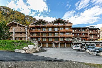 Chalet-style buildings of wood and stone with balconies, a parking lot with cars, and autumn-colored mountains under a blue, cloudy sky.