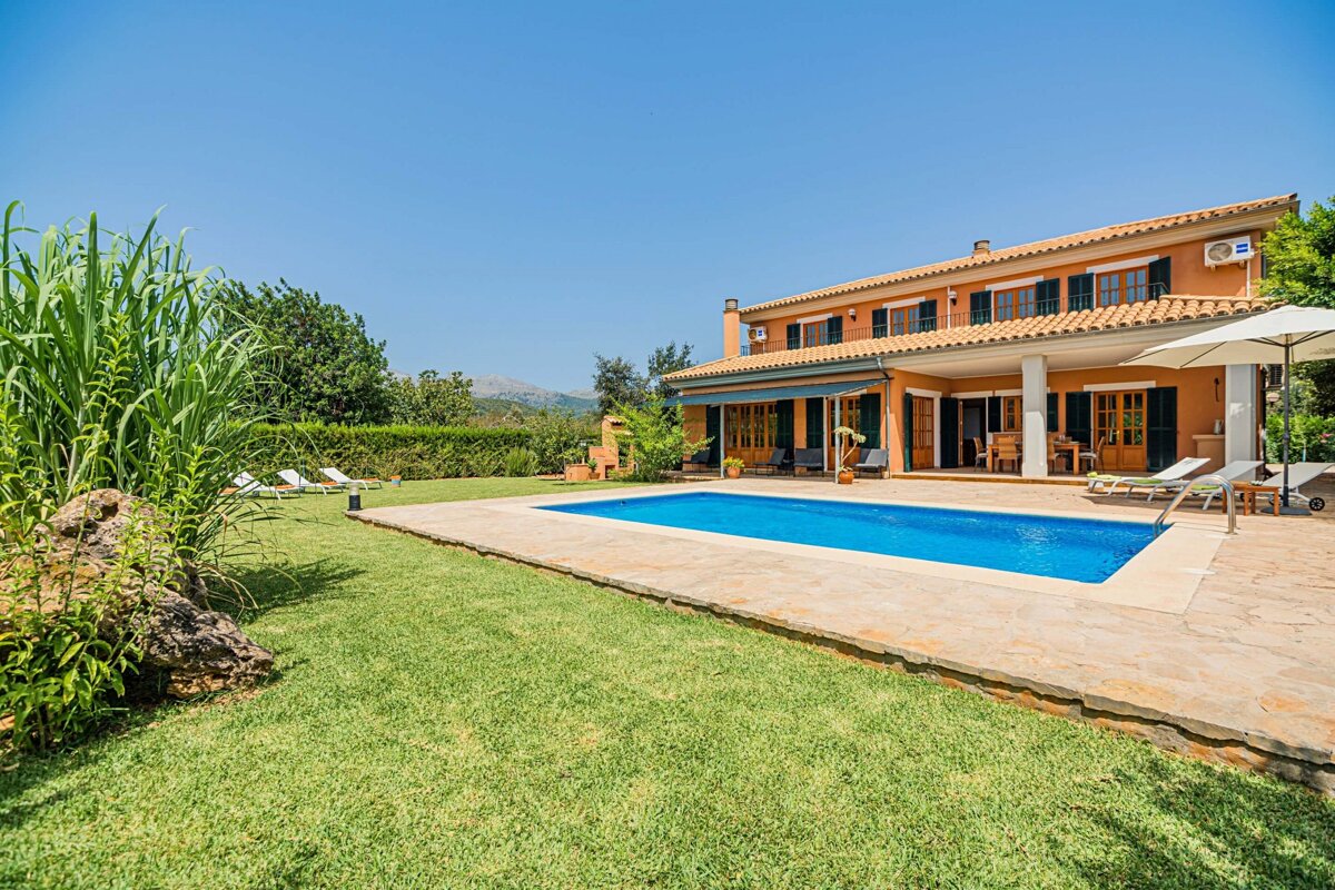 A spacious orange villa with a sparkling blue swimming pool, green lawn, and distant mountains under a clear sky, suggesting a luxurious retreat.