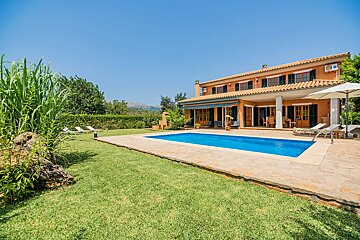 A spacious orange villa with a sparkling blue swimming pool, green lawn, and distant mountains under a clear sky, suggesting a luxurious retreat.