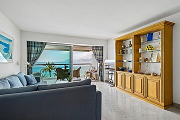 A bright living room with a large blue sectional sofa, a wooden display cabinet, and a balcony offering ocean views.
