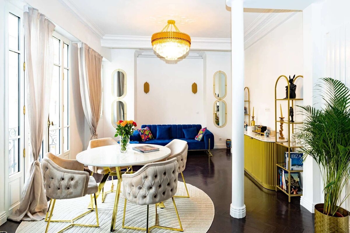 Chic living/dining room featuring a blue velvet sofa, gold chandelier, tufted chairs, a round table with flowers, and a yellow bar.