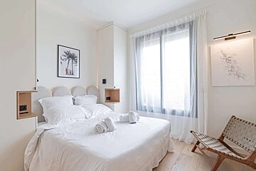 A serene, minimalist white bedroom with a plush bed, built-in nooks, a large window with sheer curtains, a palm tree print, and a woven chair.