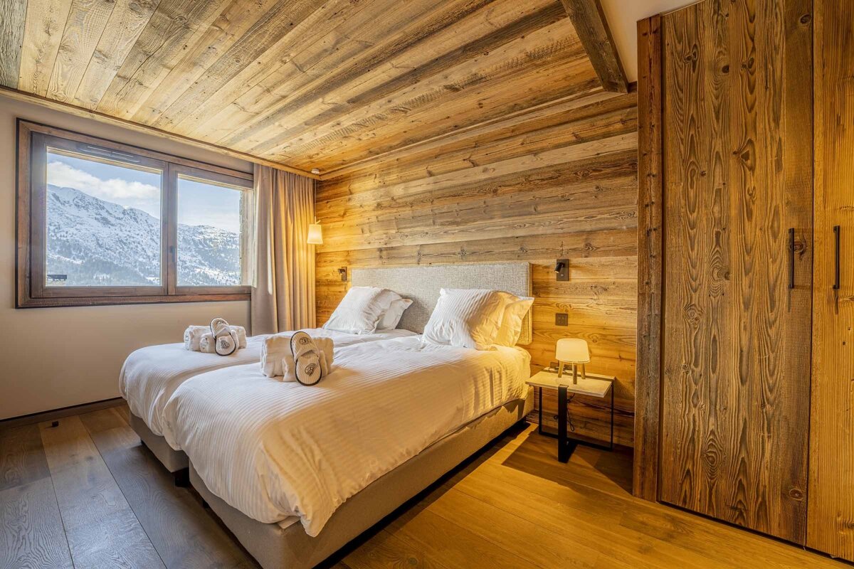 A cozy, wood-paneled chalet bedroom with two beds, white linens, and slippers. A large window offers a picturesque snowy mountain view.