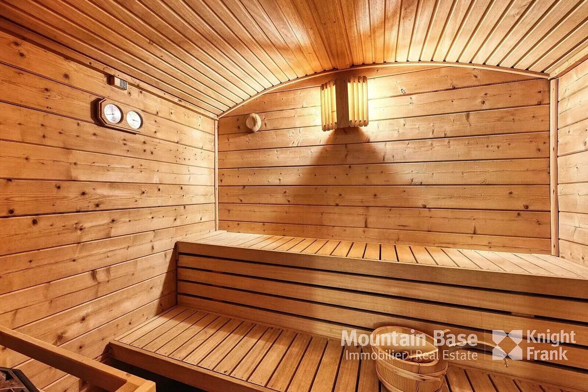 A cozy, well-lit wooden sauna featuring light wood paneling, tiered benches, a thermometer/hygrometer, and a wooden bucket with a ladle.