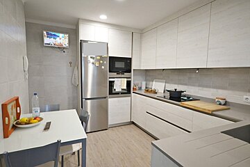 A modern kitchen with light wood cabinets, stainless steel appliances, a grey tiled backsplash, a dining table with fruit, and a TV on the wall.