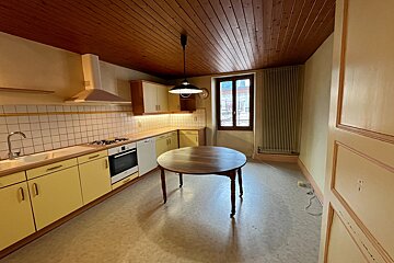An empty kitchen features yellow base cabinets, a white tiled backsplash, a dark wooden plank ceiling, and a round wooden table in the center.