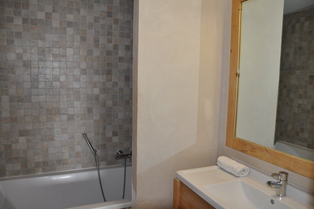 A neutral bathroom featuring a mosaic-tiled bathtub, a sink with a mirror above it, and a rolled white towel.