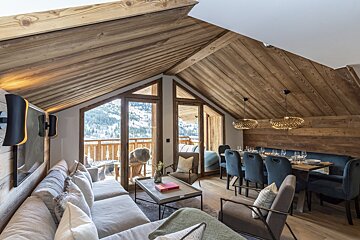 Cozy alpine chalet living and dining area with warm wood-paneling and glass doors showing snowy mountain views.