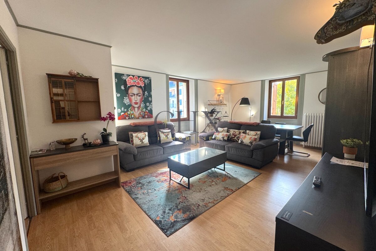 A cozy living room featuring two grey sofas, a colorful area rug, wooden floors, and a portrait of Frida Kahlo on the wall.