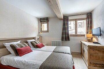 Cozy rustic twin bedroom with wood headboards, a ceiling beam, two windows with plaid curtains, and a dresser with a wall-mounted TV.