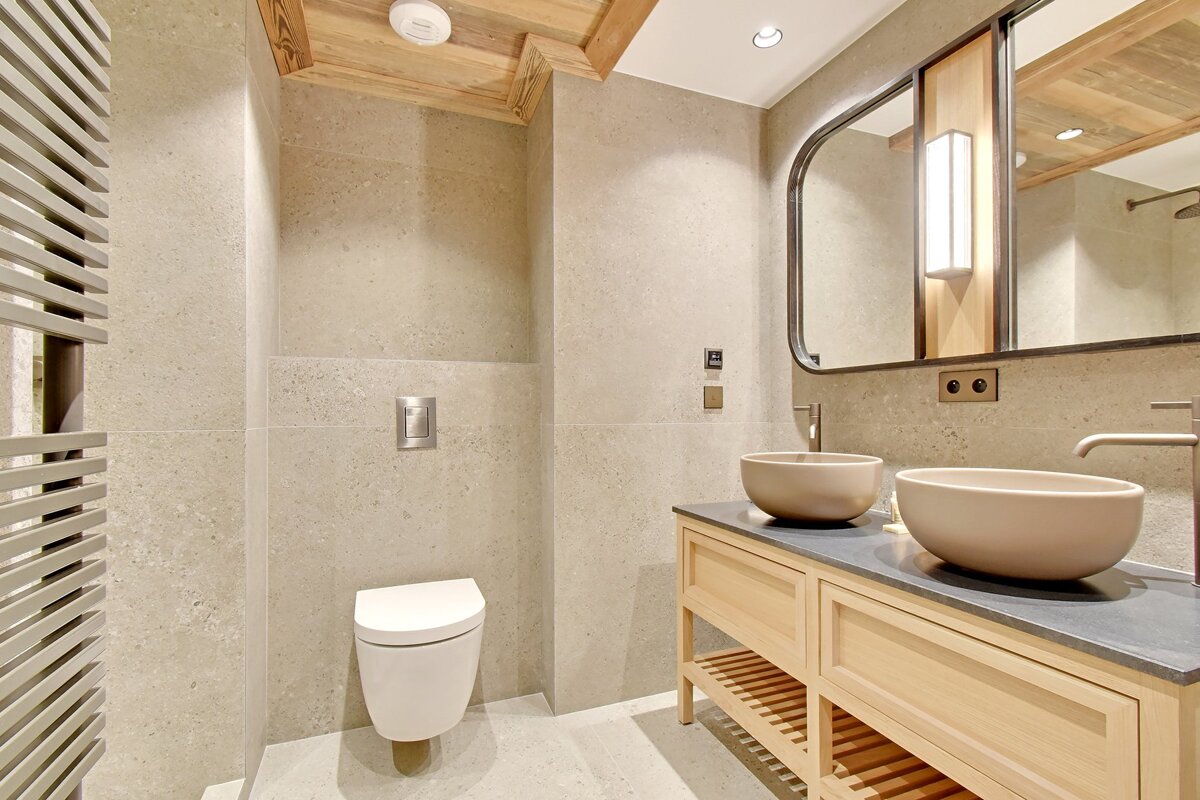 Modern bathroom featuring beige stone tiles, a wall-mounted toilet, and a light wood double vanity with two round vessel sinks.
