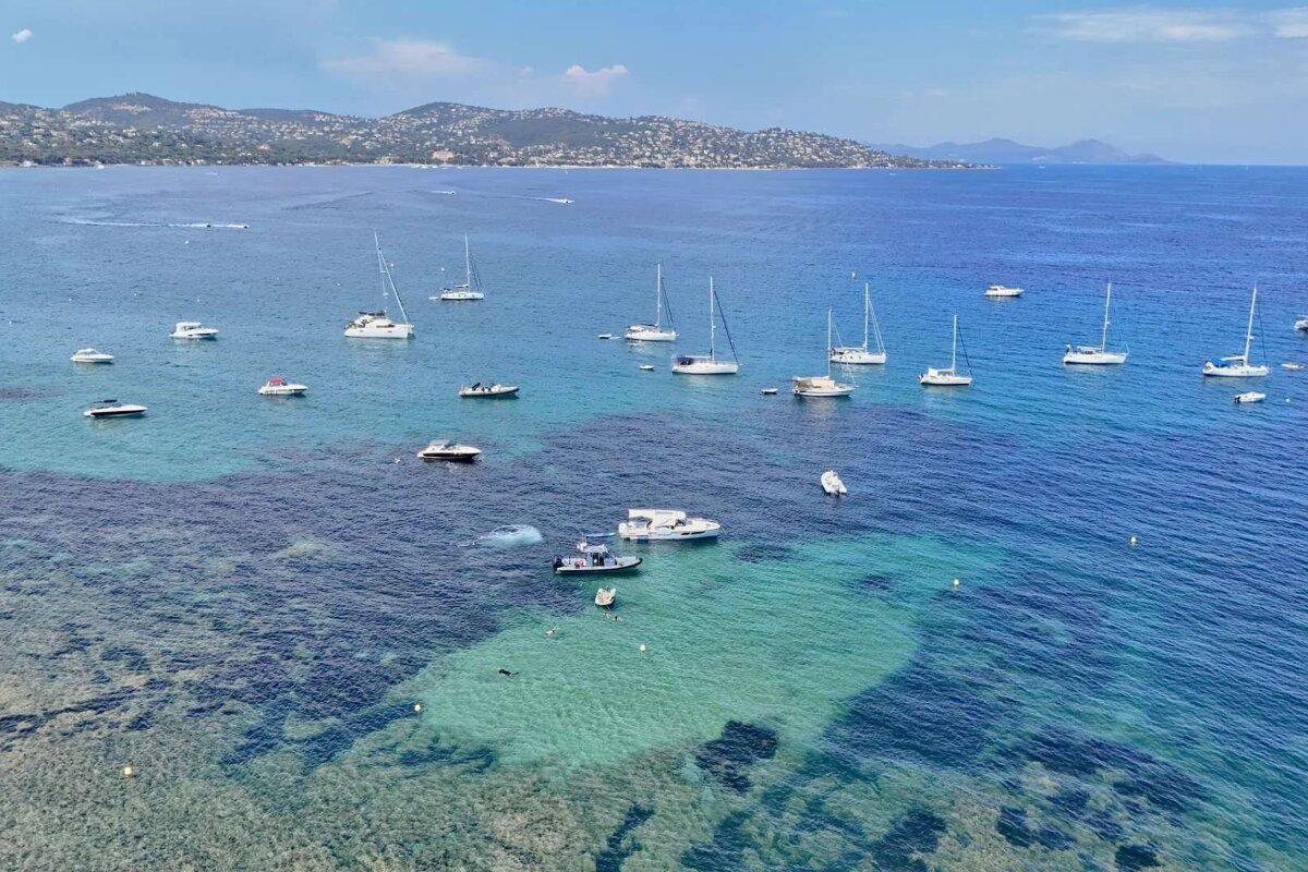 Numerous boats, including sailboats and motorboats, anchor in a clear blue sea. A lush, hilly coastline forms the backdrop under a bright sky, with shallow reefs visible underwater.