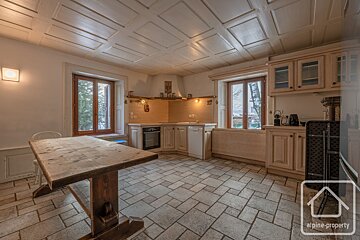 A rustic kitchen-diner with a large wooden table, tiled floor, wood-paneled ceiling, light wood cabinetry, and windows.