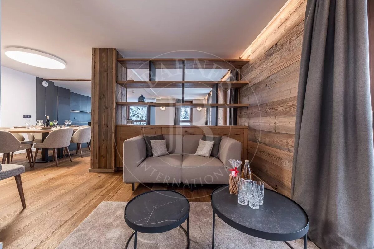 Modern open-plan living and dining area with a cozy sofa, wooden walls and shelves. A sleek kitchen is visible in the background.