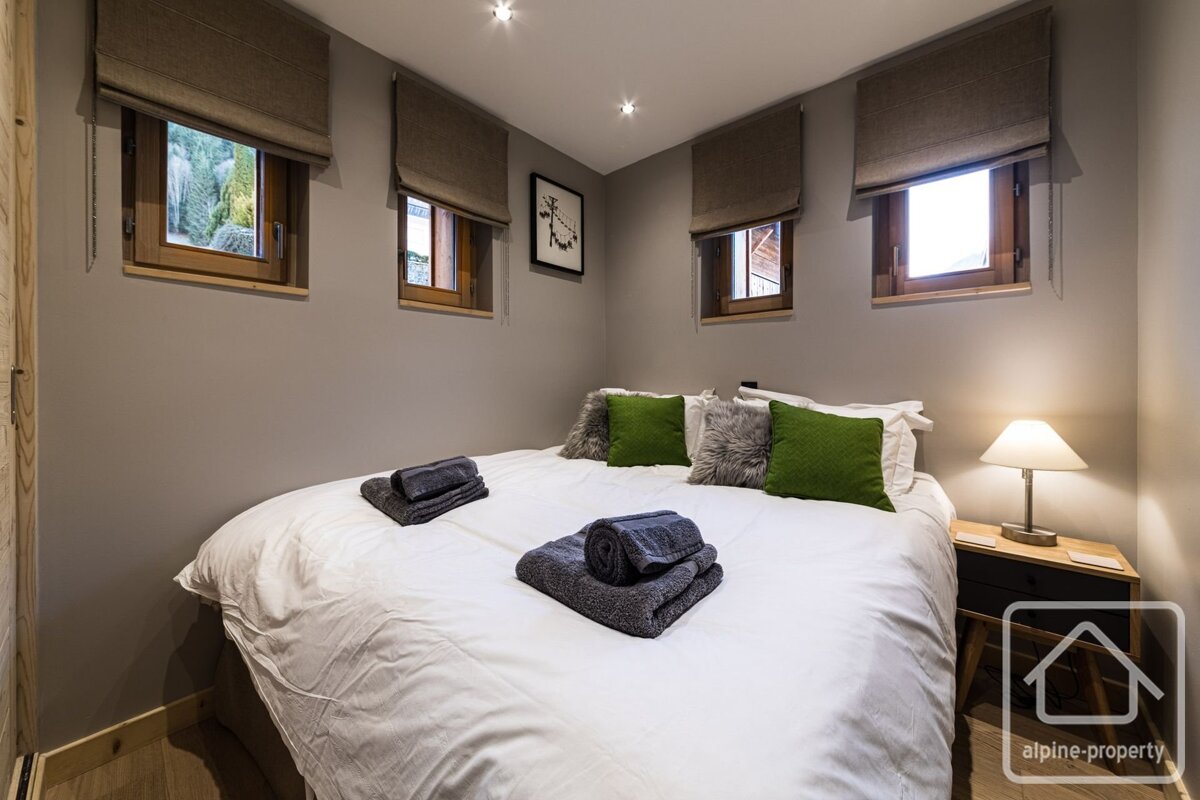 Cozy bedroom featuring a double bed with a white duvet, green pillows, folded grey towels, a nightstand with a lamp, and small windows.