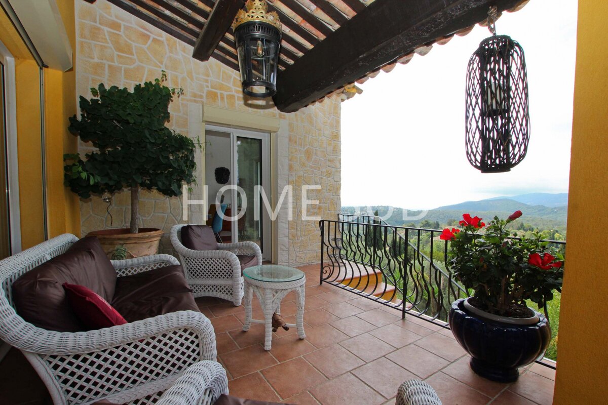 An inviting tiled balcony with white wicker furniture, potted plants, and rustic lanterns, offering a stunning panoramic view of lush green hills and mountains.