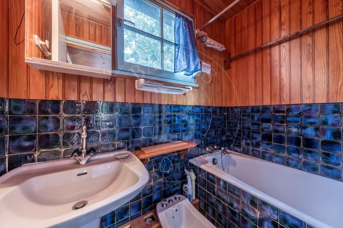 Bathroom with glossy dark blue tiles, wood-paneled walls, a white sink, bathtub, bidet, and a window.