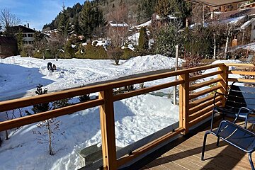 View from a wooden balcony with blue chairs, overlooking a snowy yard in an alpine village surrounded by hills and trees.