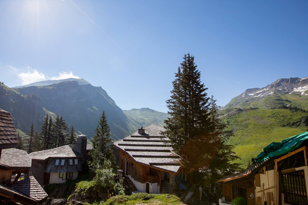 Wooden chalets nestled among pine trees in a green mountainous valley under a clear sunny sky.