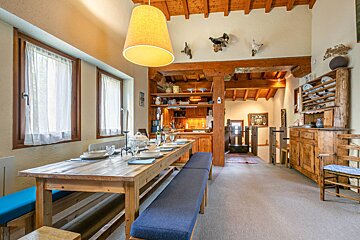 A rustic dining room with a long wooden table, blue benches, and place settings. Exposed wooden beams, a kitchen, and traditional decor.
