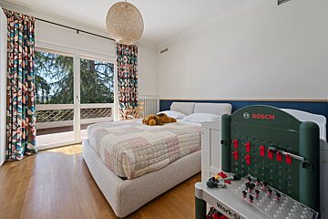 A bright child's bedroom features a bed with a quilted cover, colorful curtains, a balcony view, and a Bosch toy workbench.