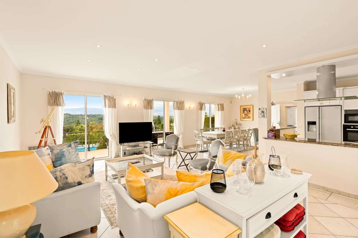 A bright, open-plan living room with yellow accents, dining area, and kitchen, featuring large windows overlooking a scenic landscape with a pool.
