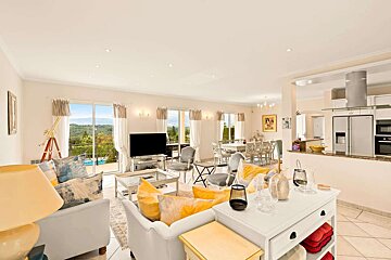 A bright, open-plan living room with yellow accents, dining area, and kitchen, featuring large windows overlooking a scenic landscape with a pool.