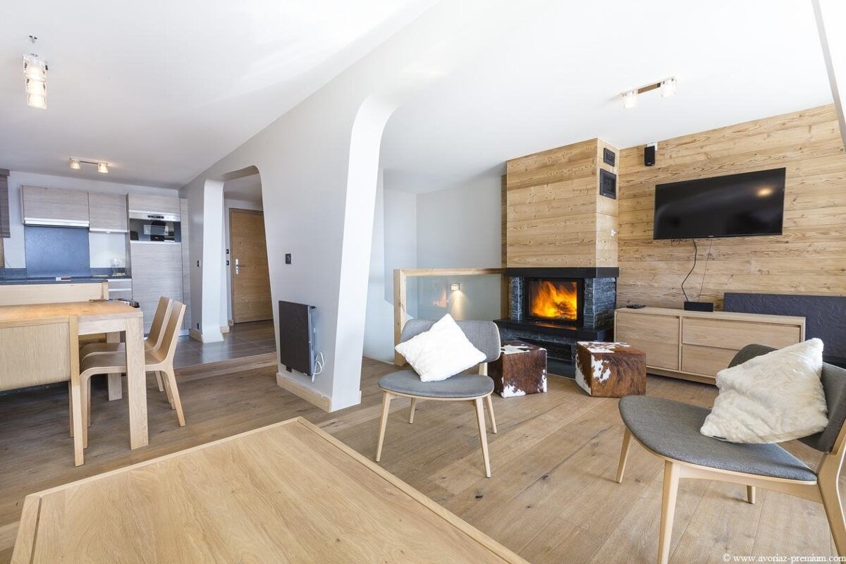Modern open-plan living room with a cozy fireplace, wood paneling, TV, and an adjacent dining/kitchen area, featuring a chic alpine aesthetic.