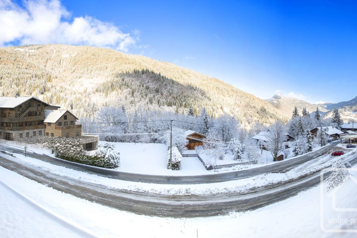 Snowy mountain village with chalets, a winding road, and forested peaks under a bright blue sky.