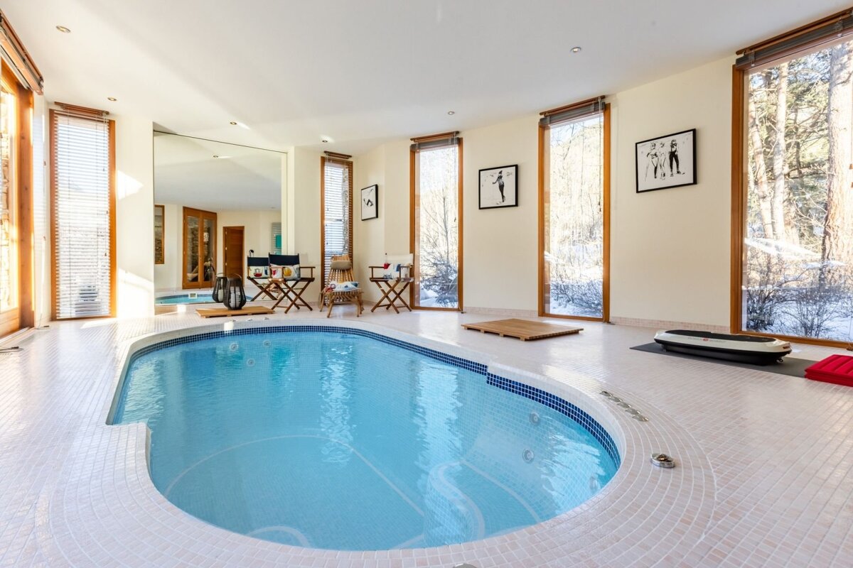 Luxurious indoor spa room with a blue oval pool, white tiles, framed wall art, and large windows showing a snowy winter landscape.