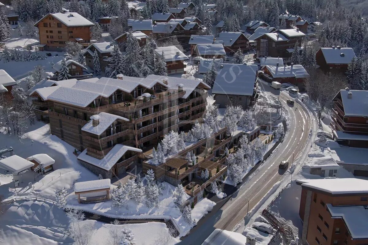 Aerial view of a snow-covered mountain village with traditional chalets, a large multi-story building, snowy trees, and a winding road.