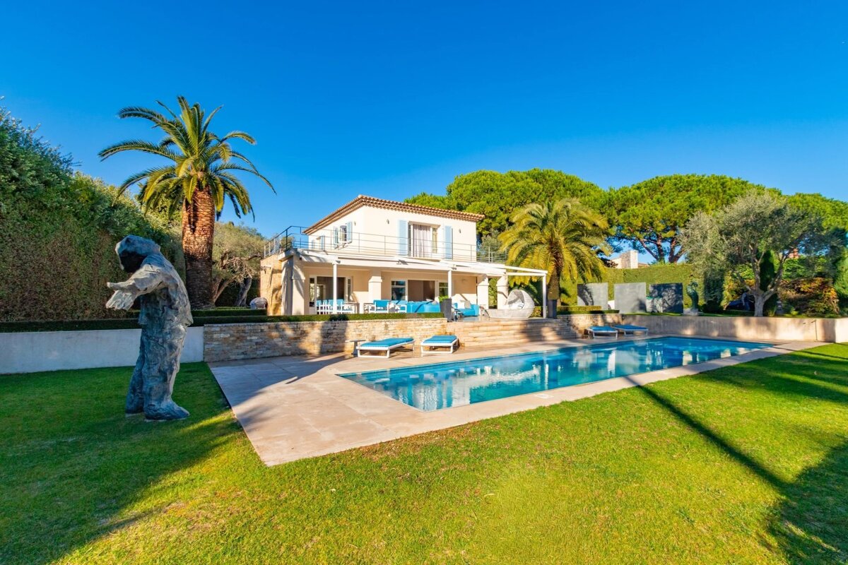 A luxurious villa with a large swimming pool, lush green lawn, palm trees, outdoor seating, and a sculpture, under a clear blue sky.