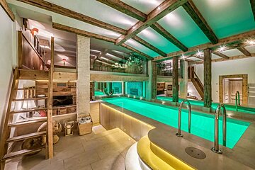 Luxurious indoor pool glows green in a rustic-modern space with exposed wood beams, multi-level design, and warm ambient lighting.