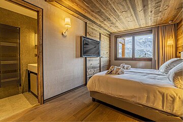 Modern alpine chalet bedroom featuring rustic wood paneling, a large bed with a mountain view, and an adjacent bathroom.