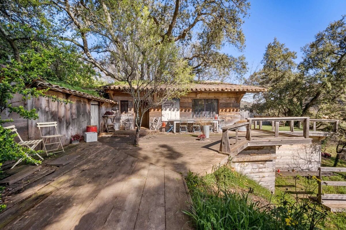 A rustic wooden house with a green roof and a large wooden deck/patio is nestled among lush trees. Outdoor furniture suggests a natural, relaxed setting.