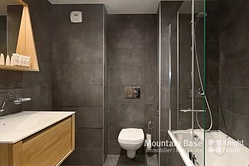 Modern bathroom with dark grey tiled walls, a wooden vanity, toilet, and a bathtub with a clear shower screen. Features toiletries and a minimalist design.