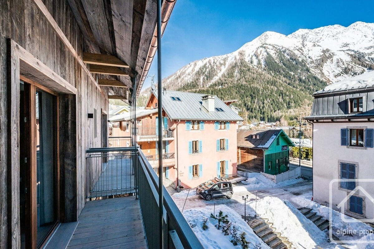 A wooden balcony offers a snowy alpine village view with colorful homes, a car, and majestic snow-capped mountains under a clear blue sky.